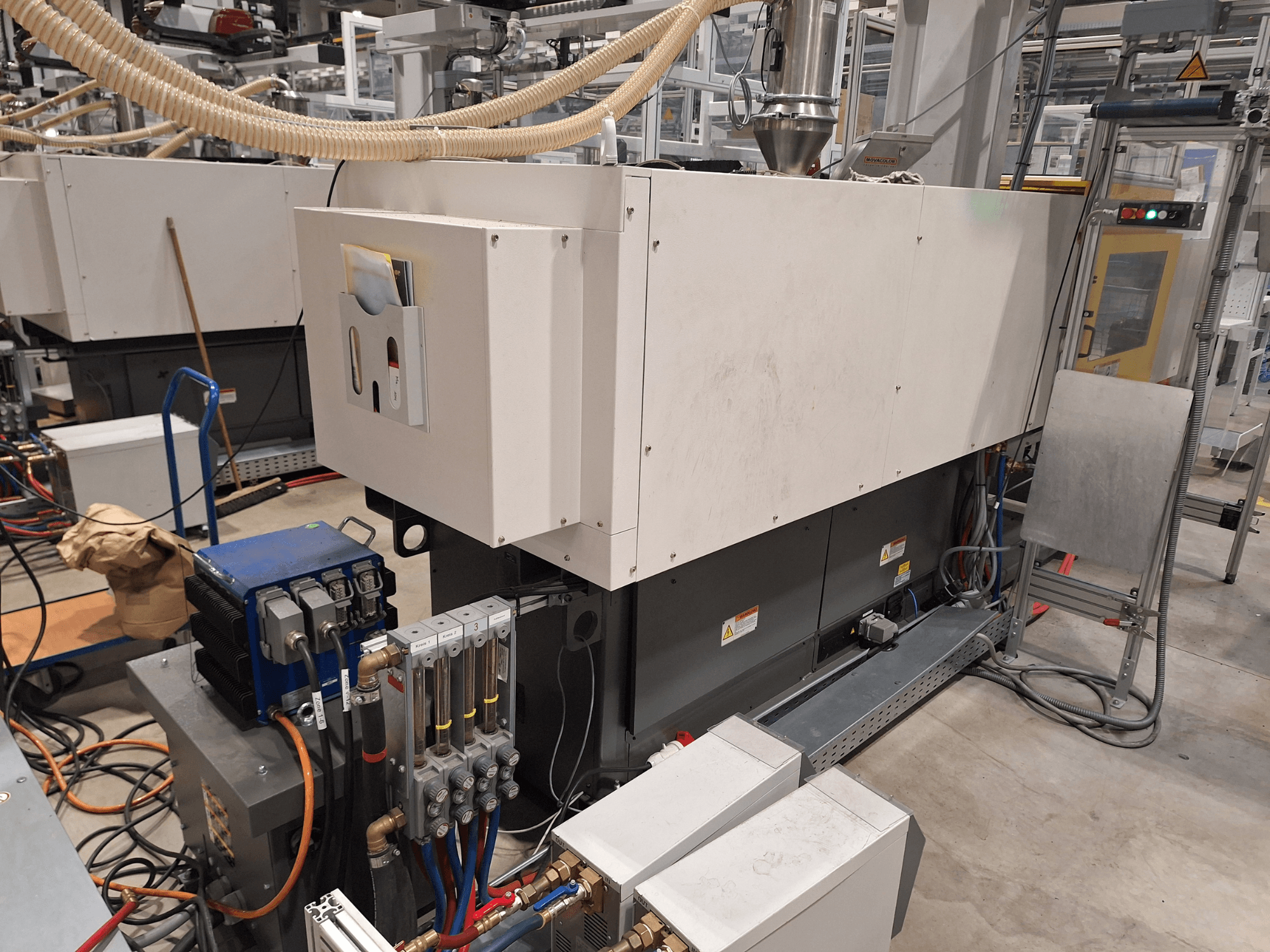 Ipari berendezések gyári környezetben; egy nagy gépet tartalmaz vezérlőpanellel, tömlőkkel és elektromos csatlakozókkal, oldalnézet.