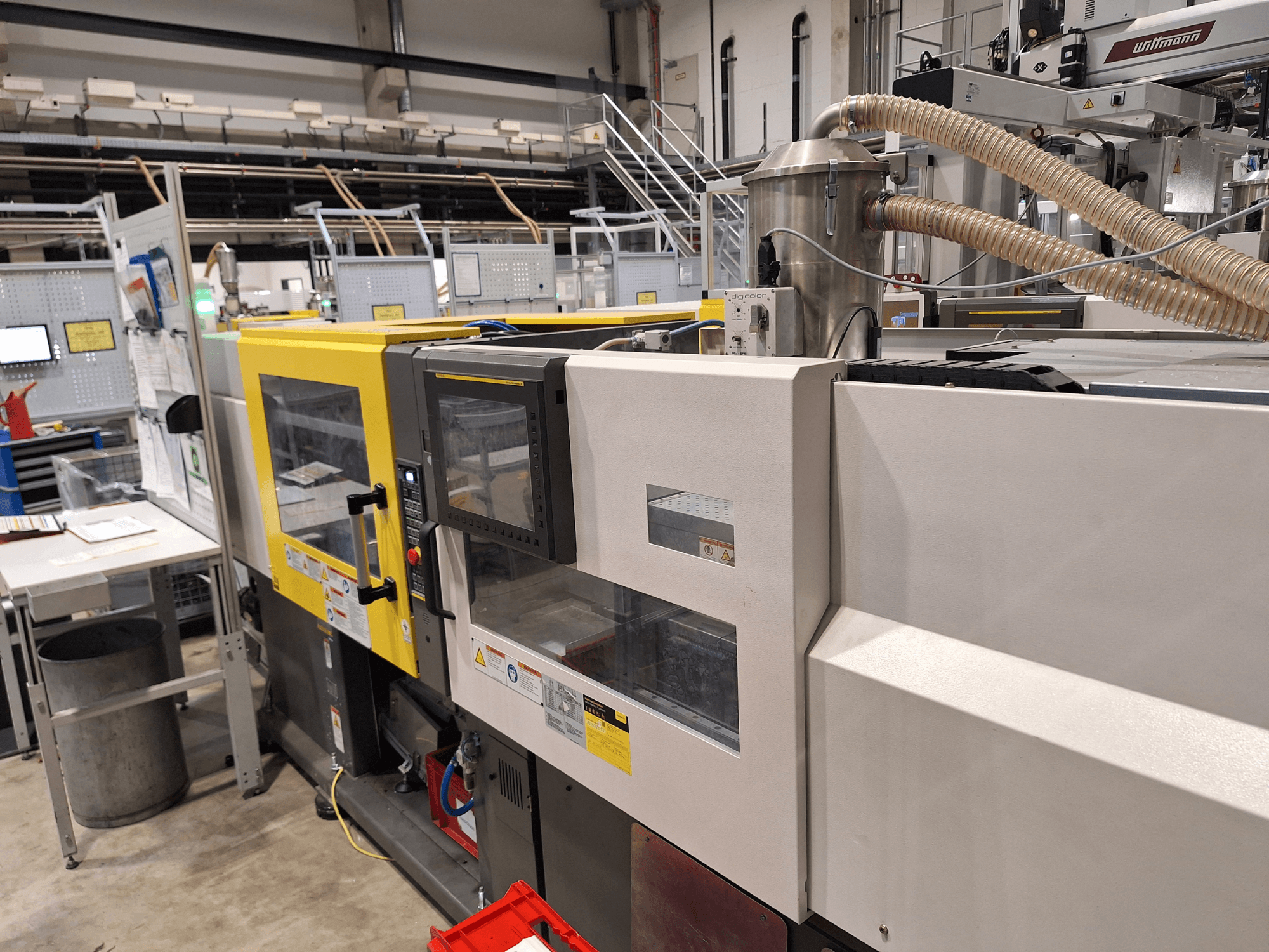 Egy többszínű, sárga és szürke részeket tartalmazó fröccsöntőgép elülső nézete, vezérlőpanellel és kipufogórendszerrel.