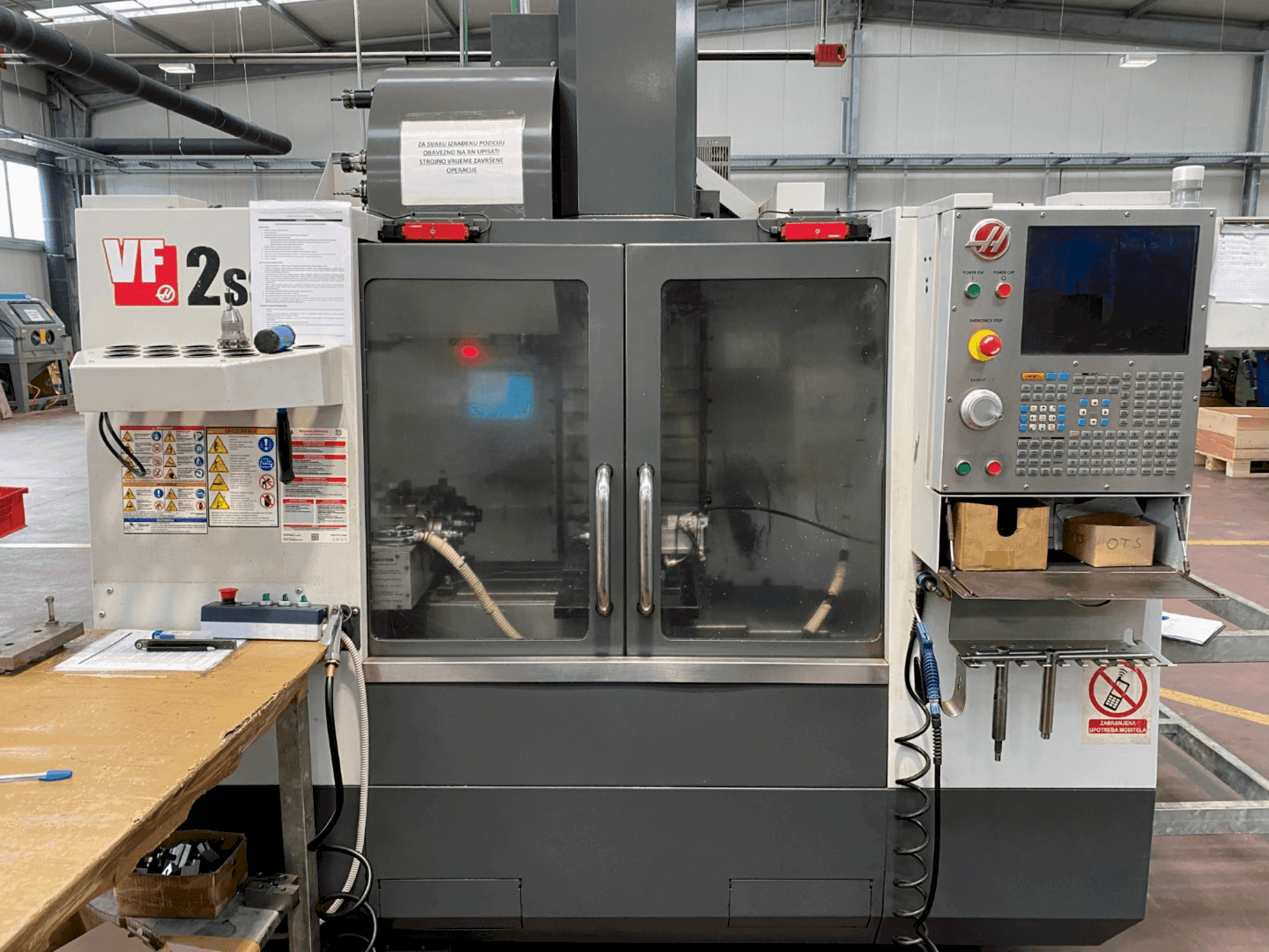 Haas VF-2SS megmunkálóközpont elölnézetben, vezérlőpanel, hűtőfolyadék-tömlő és biztonsági címkék az oldalán.